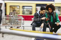 Man and woman looking at phone while sitting on a bench on a train platform.