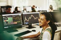 Woman smiling while sitting behind her desk, screens displaying financial information.