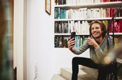 Smiling man sitting on stairs, making a selfie. bookshelf in background