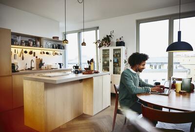 Smiling man sitting at his dining table working on his laptop. Kitchen in the background.